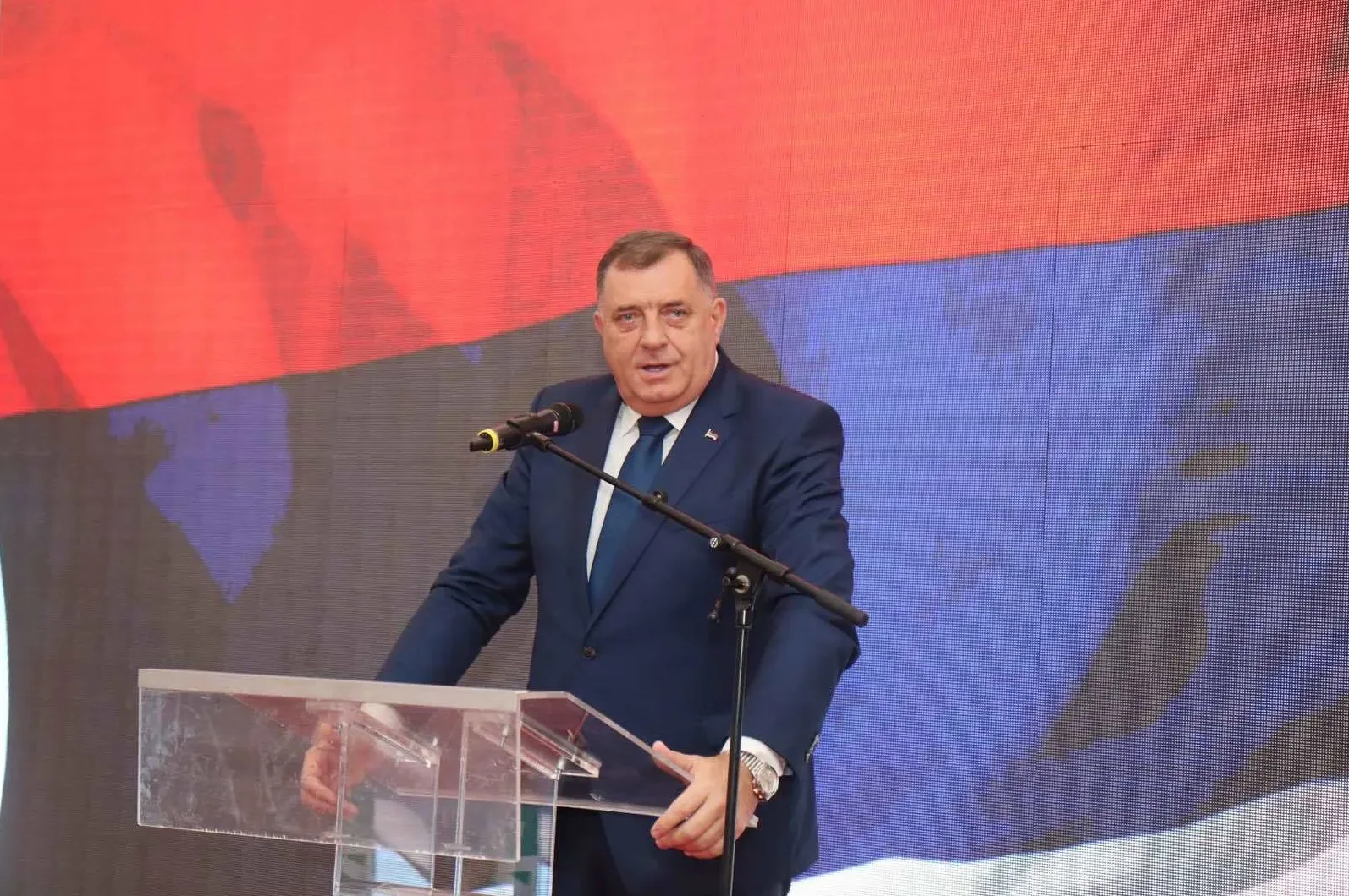 Milorad Dodik, predsjednik SNSD-a i Republike Srpske, stoji iza podijuma i drži govor ispred zastave Republike Srpske.