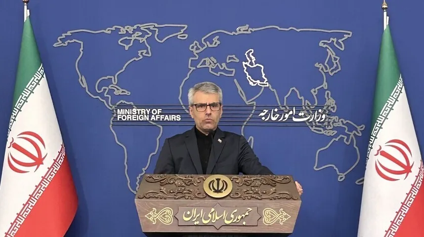 Portret muškarca u tamnom odijelu i crnoj košulji koji stoji iza drvenog podijuma na kojem je ispisano 'Republika Iran' na perzijskom jeziku i grb Irana, a u pozadini se nalazi mapa svijeta i iranske zastave.
