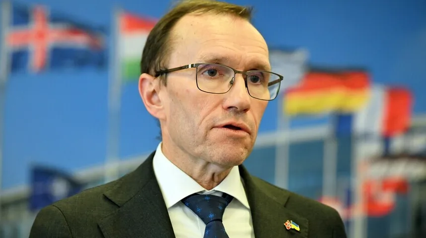 Portret muškarca u tamnom odijelu i plavoj kravati s uzorkom, nosi naočale, u pozadini se vide zastave zemalja.