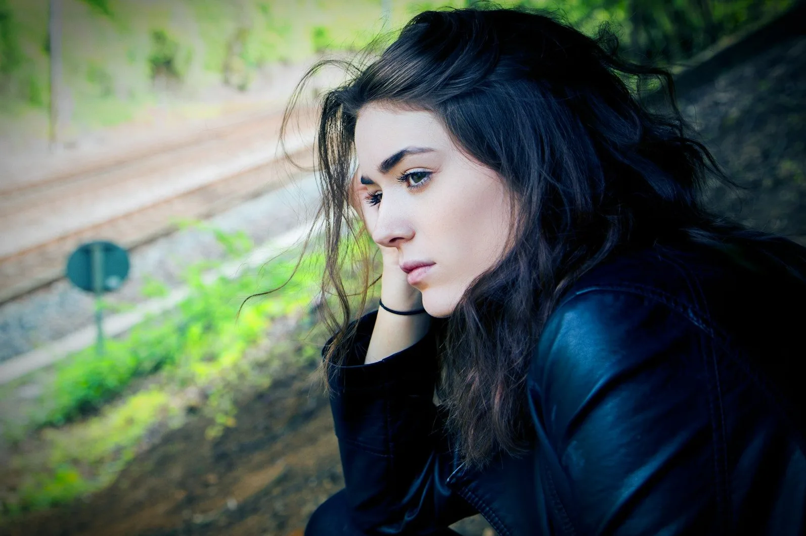 Portret mlade djevojke tamne kose i ozbiljnog pogleda, odjevene u kožnu jaknu, koja sjedi pored željezničke pruge i zelenog rastinja, duboko zadubljena u misli.