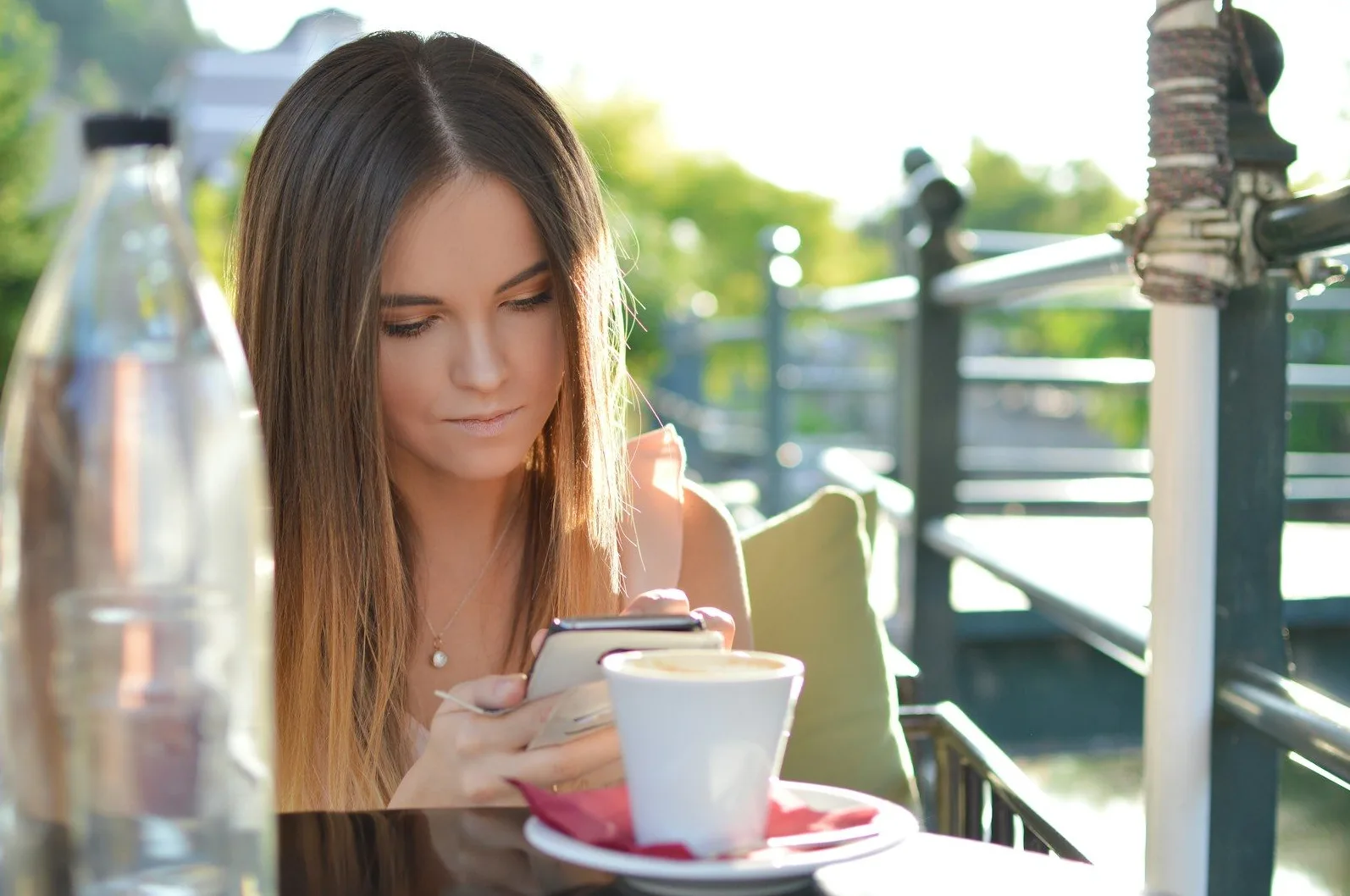 Mlada žena sjedi vani i koristi mobilni telefon, s šoljicom kafe pored.