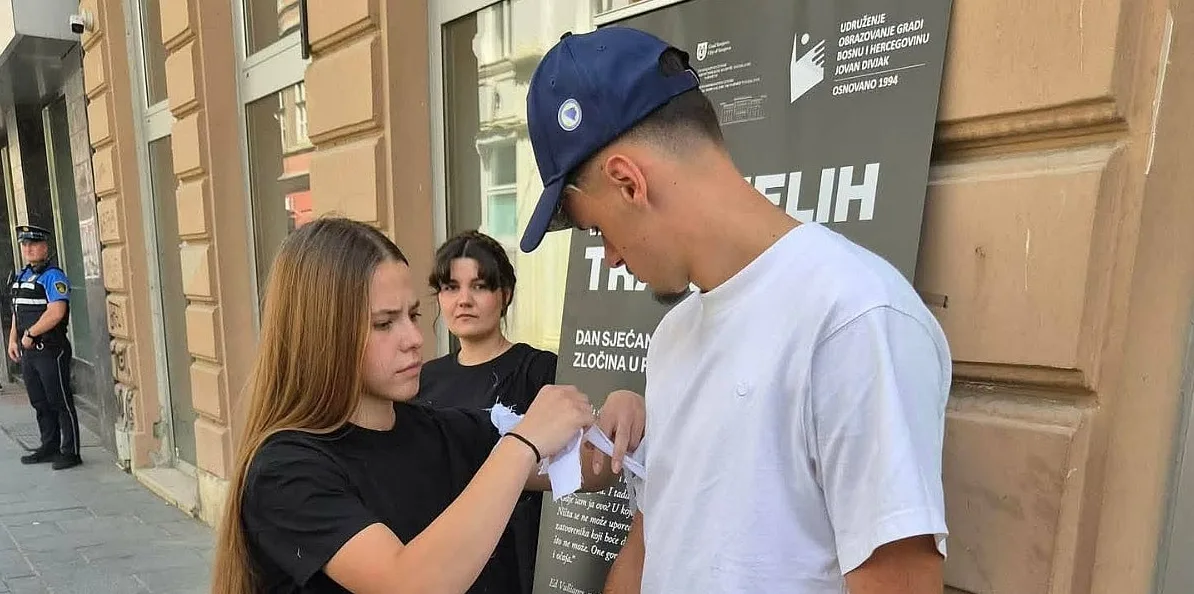 Djevojka i mladić razmjenjuju papirnu traku ispred zgrade sa natpisom Udruženje "Obrazovanje gradi Bosnu i Hercegovinu" Jovan Divjak, osnovano 1994.