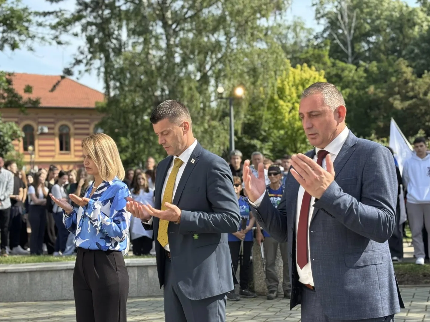 Dvoje ljudi u svečanim odijelima, muškarac i žena, sa podignutim rukama u molitvi, okruženi gomilom ljudi na otvorenom.