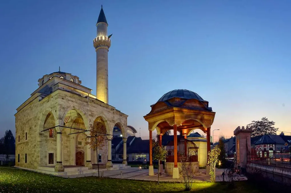 Džamija i drvena šadrvan fontana osvijetljene noću sa plavim nebom u pozadini.