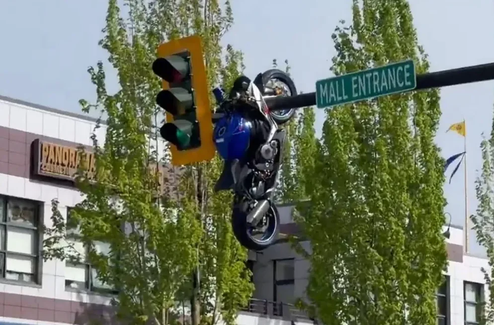 Motociklista izvodi akrobaciju na motociklu pored semafora i znaka za ulaz u tržni centar.