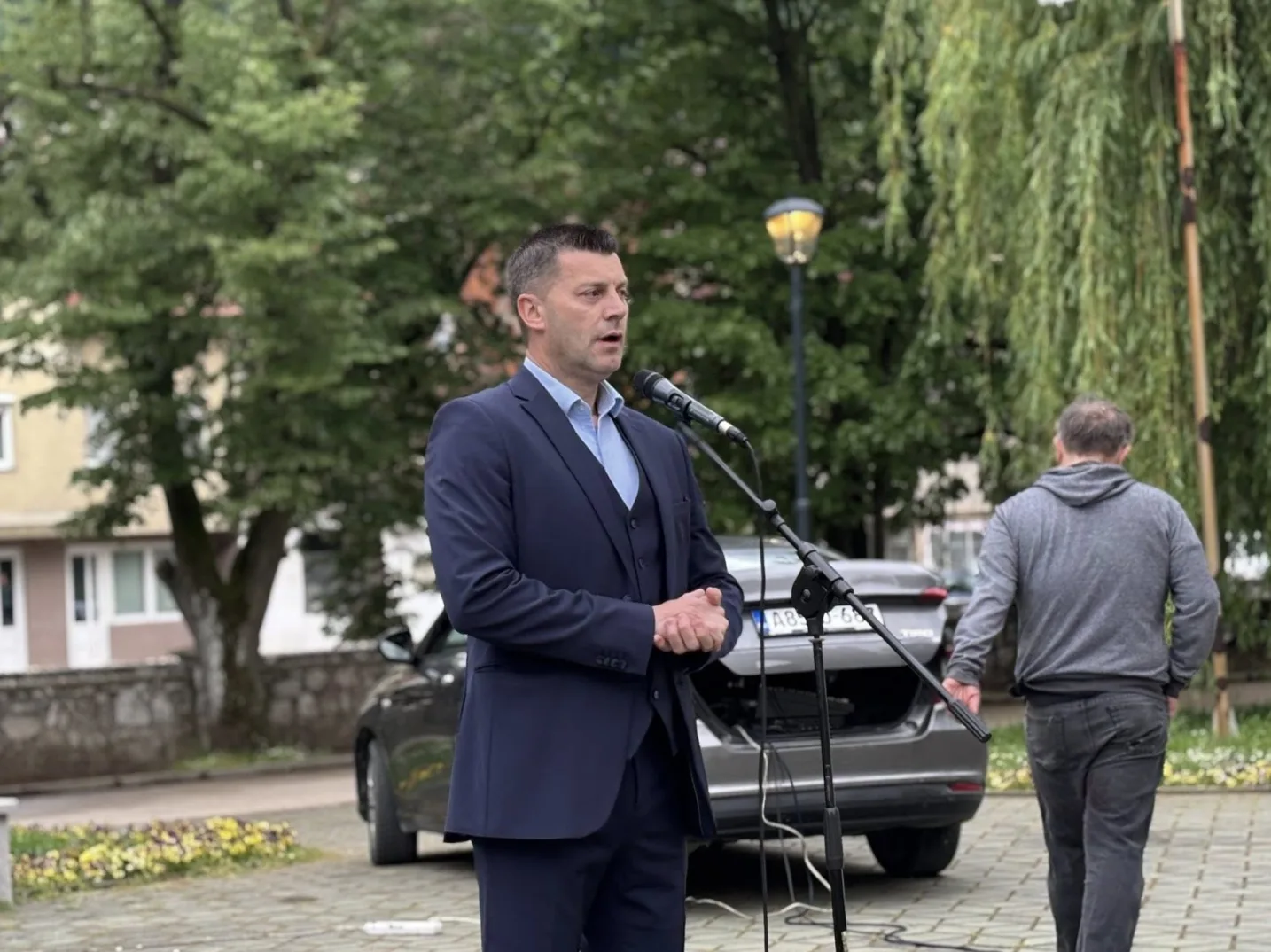 Političar u tamnom odijelu govori ispred mikrofona, sa automobilom u pozadini.