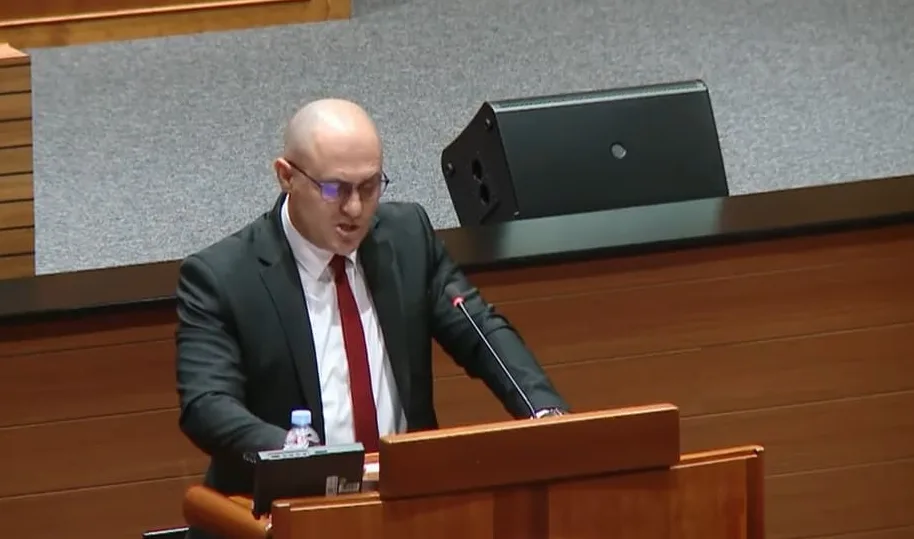 Muškarac u odijelu sa crvenom kravatom i naočalama govori za govornicom u sali za sjednice.