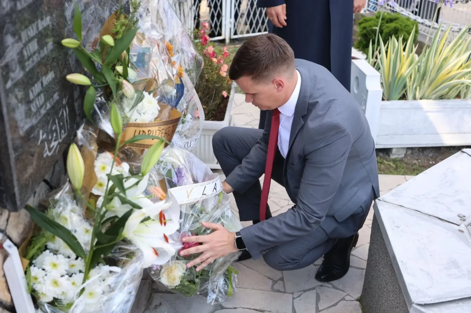 Muškarac u sivom odijelu i crvenoj kravati čuči pored nadgrobnog spomenika i polaže cvijeće. Iza njega se vide ostali ljudi i cvjetni aranžmani.