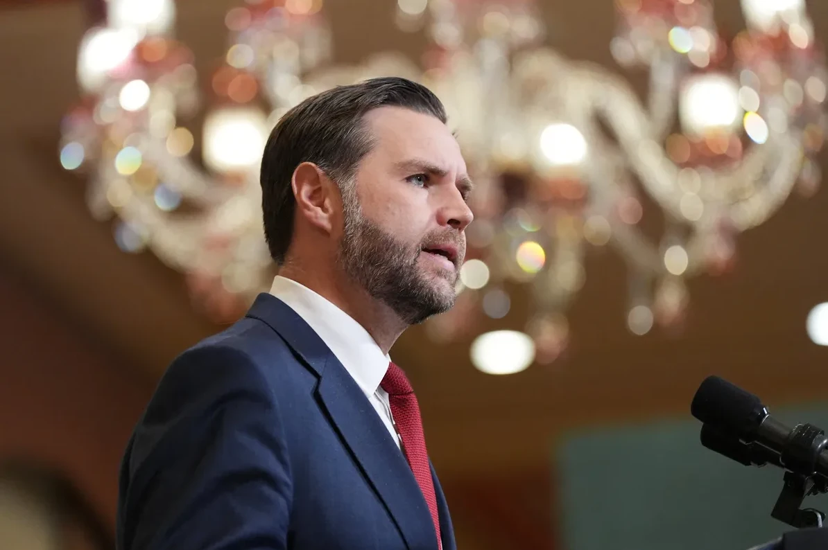 Muškarac sa bradom u tamnoplavom odijelu i crvenoj kravati govori za govornički mikrofon.