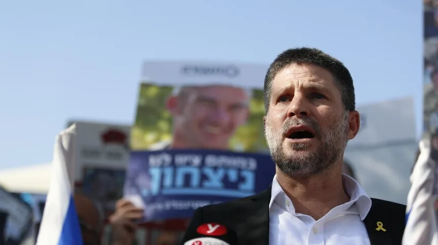 Političar sa bradom govori na skupu, držeći žutu vrpcu prikačenu na reveru, dok se u pozadini vide plakati sa slikama osoba.