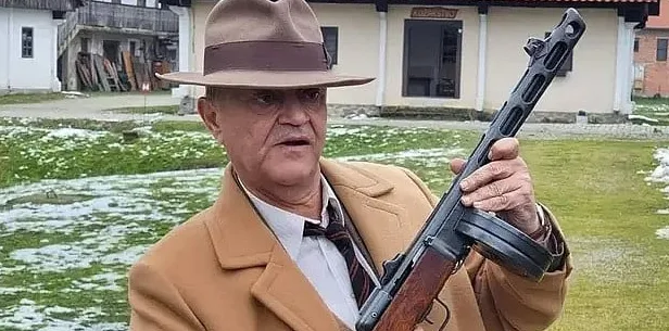 Stariji muškarac u bež kaputu i šeširu drži automatsku pušku.