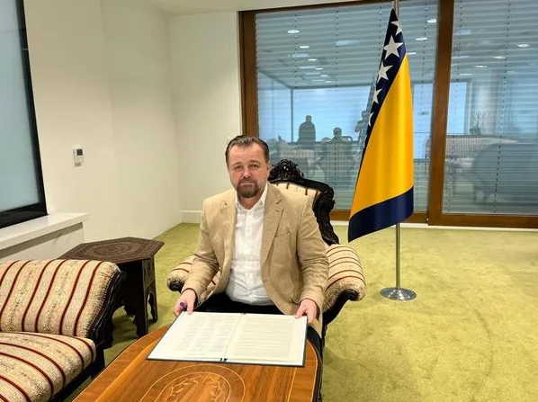 Političar u elegantnom odijelu sjedi u stolici pored zastave Bosne i Hercegovine.