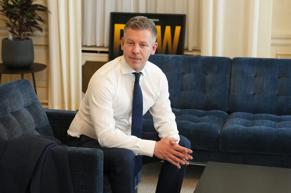 Muškarac u bijeloj košulji i tamnoplavoj kravati sjedi na tamnoplavoj garnituri sa rukama ukrštenim.