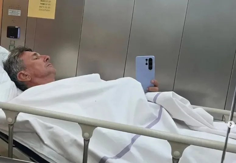 Muškarac srednje životne dobi spava u bolničkom krevetu, pokriven bijelim pokrivačem, držeći svijetloplavi mobilni telefon u desnoj ruci.
