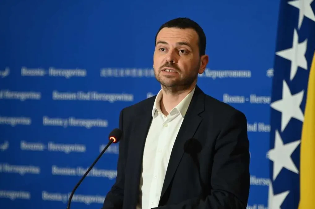 Portret muškarca u tamnom odijelu i bijeloj košulji, koji govori za govornicom. U pozadini se vidi plavi zastor sa ispisanim nazivom države Bosna i Hercegovina na više jezika, te zastava Bosne i Hercegovine.