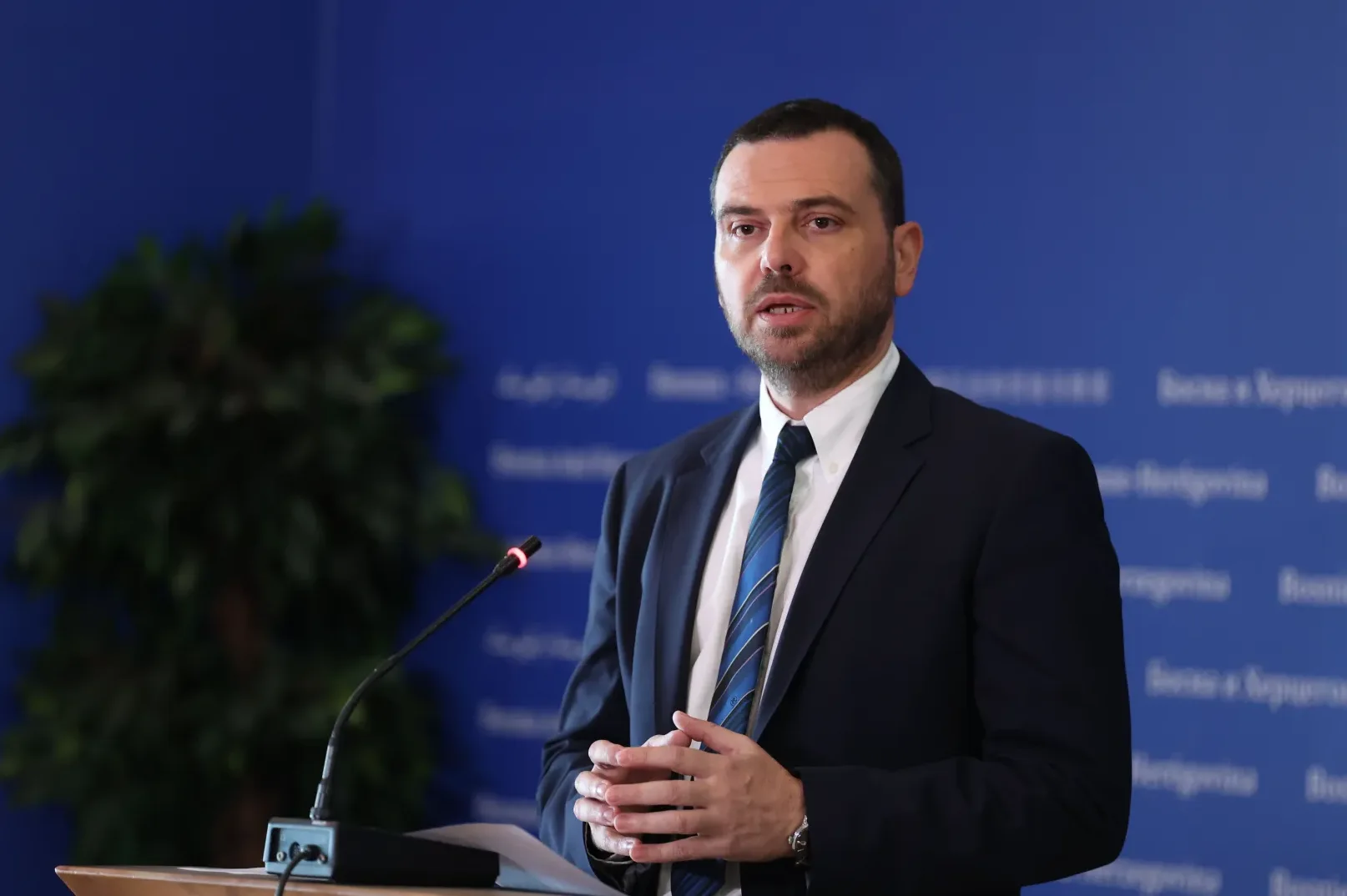 Političar ili zvaničnik u tamnom odijelu i plavoj kravati govori za govornicom ispred plave pozadine sa logom.