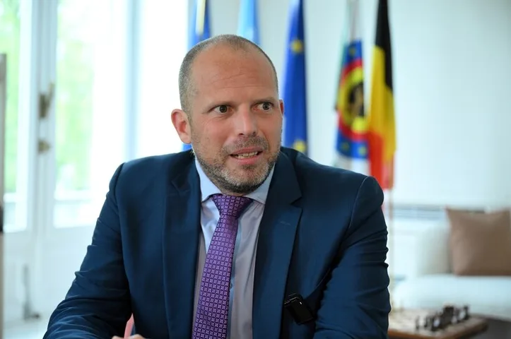 Muškarac u tamnoplavom odijelu i ljubičastoj kravati sa sitnim uzorkom, sjedi i govori. Na poleđini se vide zastave Evropske unije i Belgije.