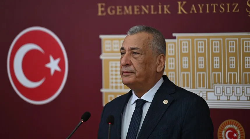 Portret starijeg muškarca u tamnom odijelu i bijeloj košulji sa tamnom kravatom, kako govori za govornicom ispred zastave Turske i stilizovane slike zgrade parlamenta.