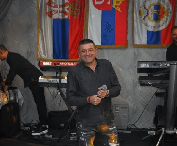 Čovjek sa mikrofonom u ruci, nasmijan, stoji ispred nekoliko zastava.