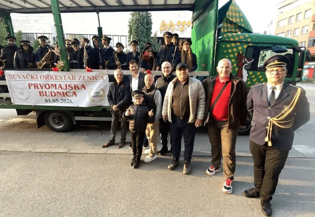 Grupa ljudi u uniformama sa duvačkim instrumentima i dječačić u kapici duvačkog orkestra stoje ispred kamiona na kojem piše "Duvacki orkestar Zenica - Prvomajska Budnica 01.05.2026. 100 godina".