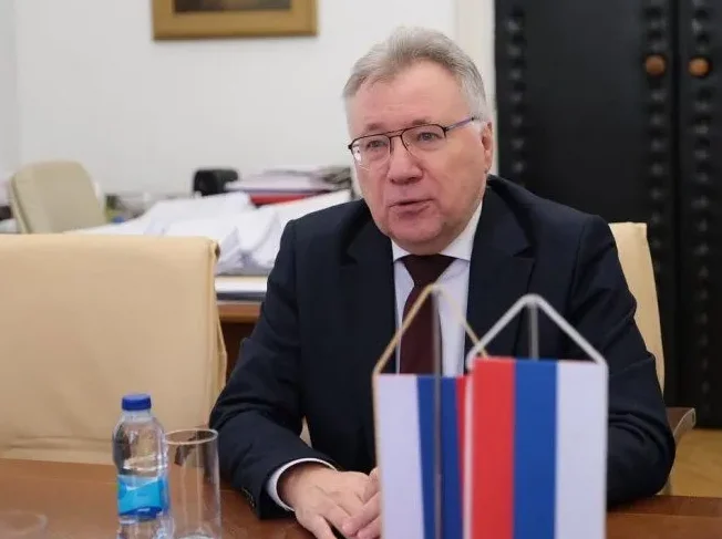 Čovjek u tamnom odijelu, bijeloj košulji i tamnom bordo kravati, sa naočalama, sjedi za stolom ispred dvije zastavice Rusije.