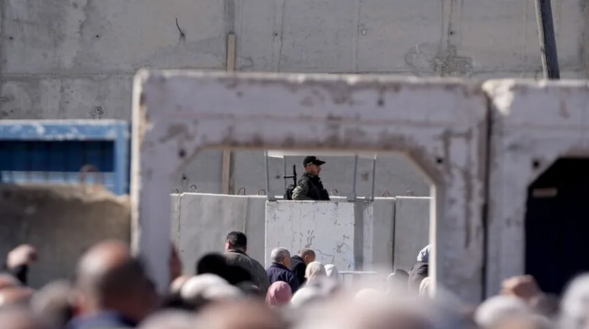 Osoba u uniformi i sa naoružanjem nadgleda grupu ljudi iza betonskih prepreka.