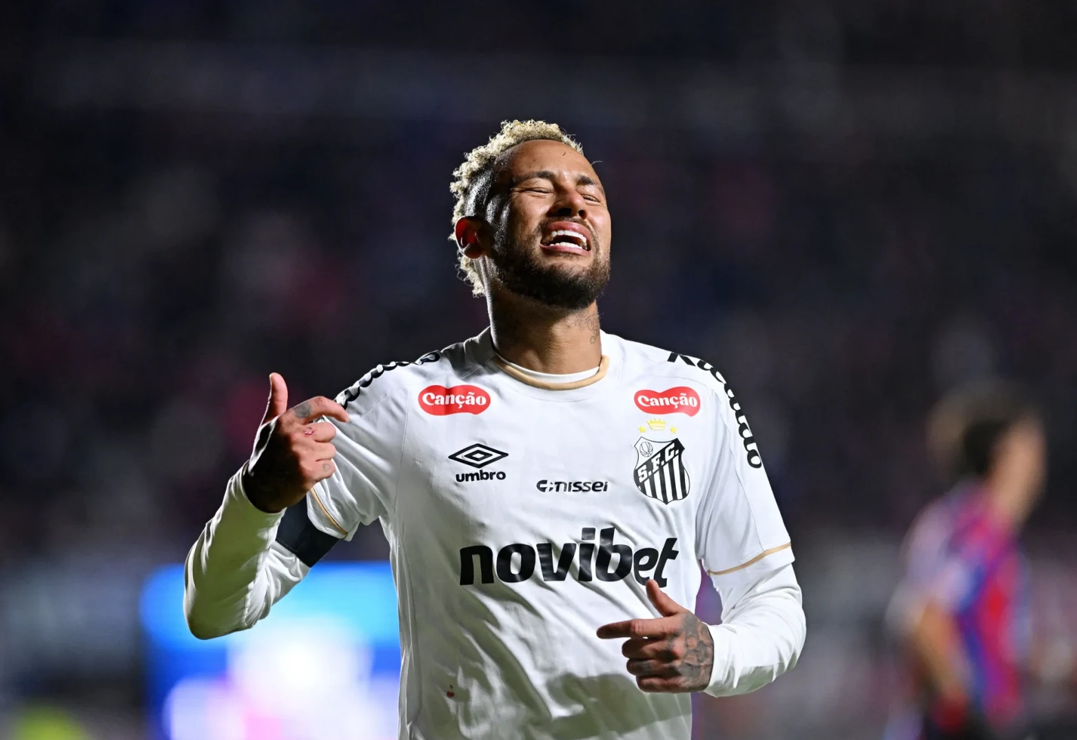 Fudbaler Neymar Jr. u bijelom dresu sa logom Sanca, sa podignutim palcem i izrazom lica koji ukazuje na bol ili razočarenje.