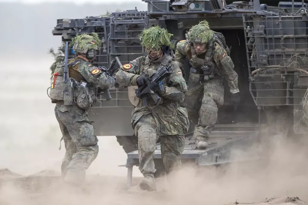 Njemački vojnici u punoj borbenoj opremi, sa kamuflažnim uniformama i granjem na kacigama, iskrcavaju se iz oklopnog vozila.