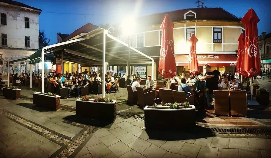 Fotografija prikazuje spoljašnji prostor kavane u večernjim satima, sa brojnim gostima koji sjede za stolovima pod crvenim suncobranima.