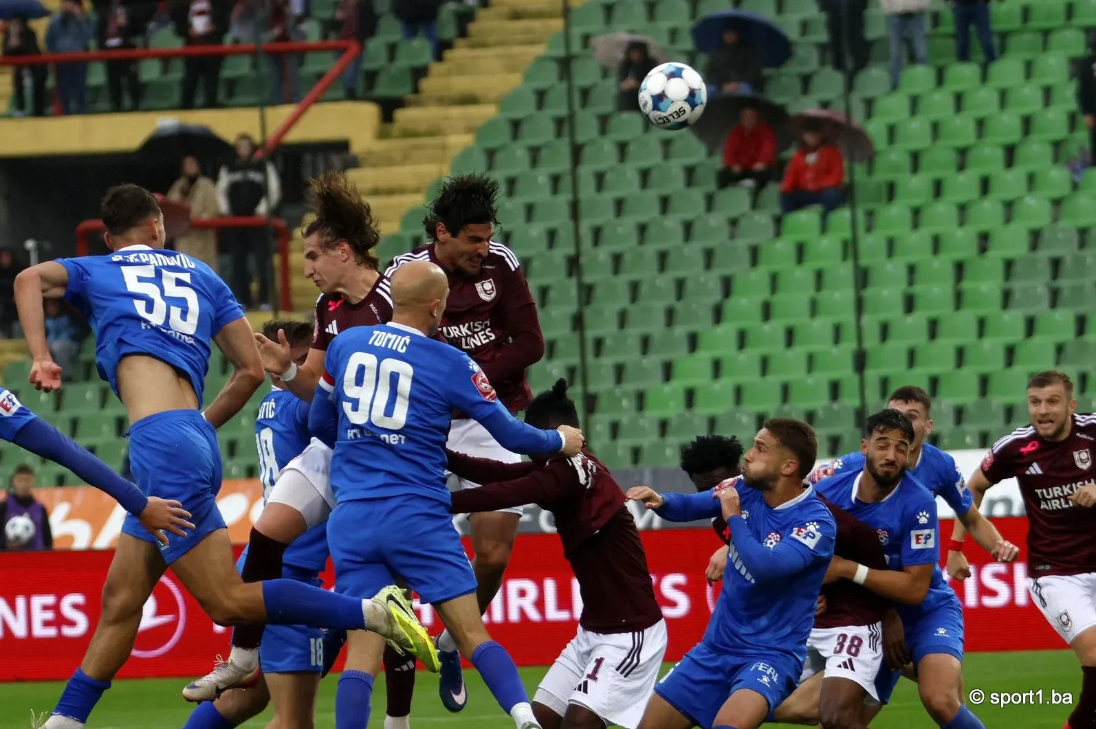 Fudbaleri u plavim i bordo dresovima bore se za loptu glavom na fudbalskom stadionu, dok publika sjedi na tribinama.