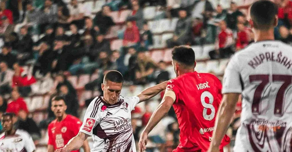 Dva fudbalera u akciji na stadionu, jedan u bijelom dresu sa natpisom "Turkish Airlines" i brojem 77, dok je drugi u crvenom dresu sa brojem 8.