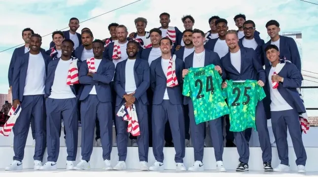 Grupa fudbalera u plavim odijelima i bijelim majicama pozira sa američkim zastavama i dresovima.