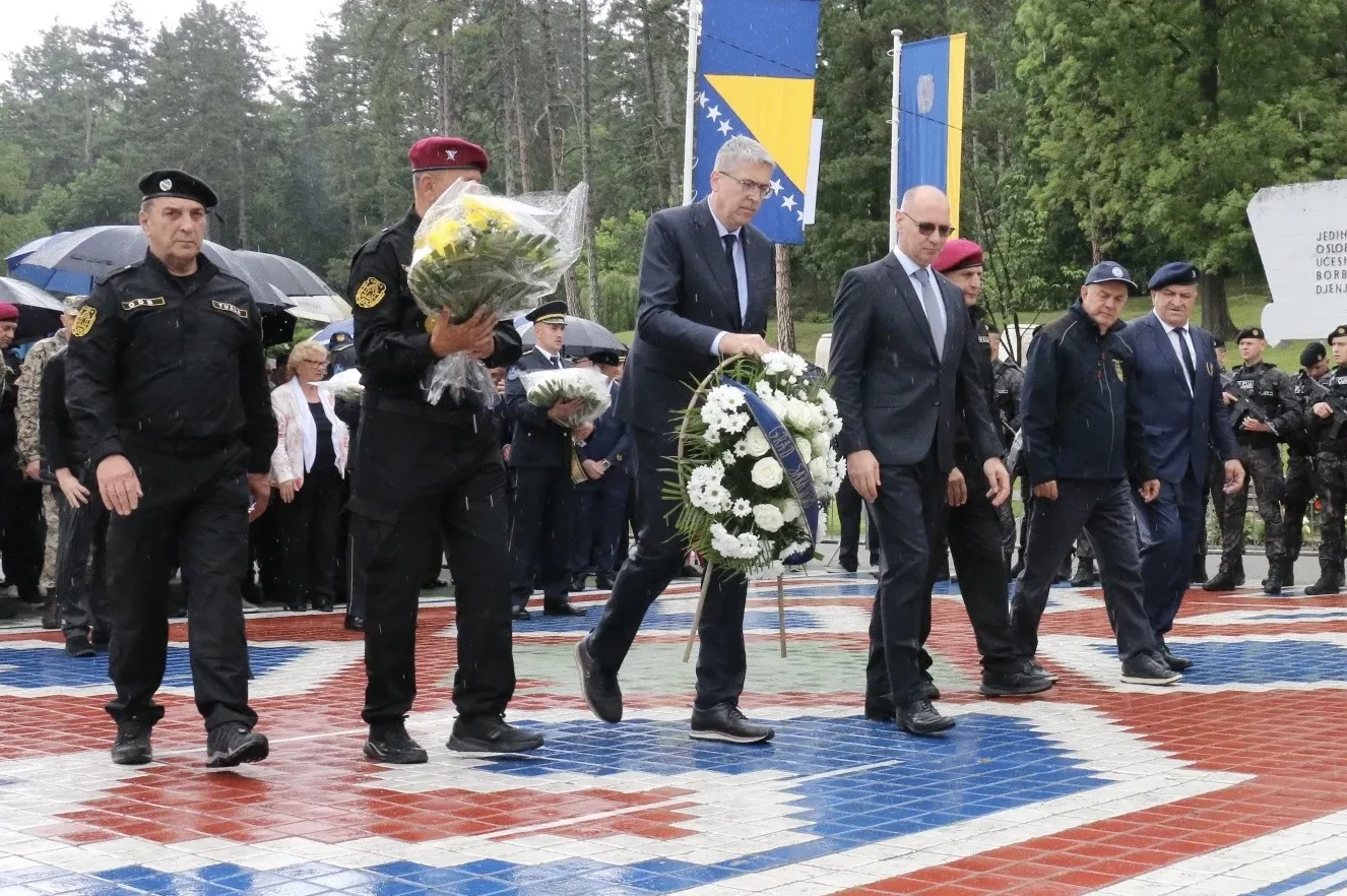 Ljudi u uniformama i odijelima nose cvjetne vijence ispred zastava Bosne i Hercegovine.