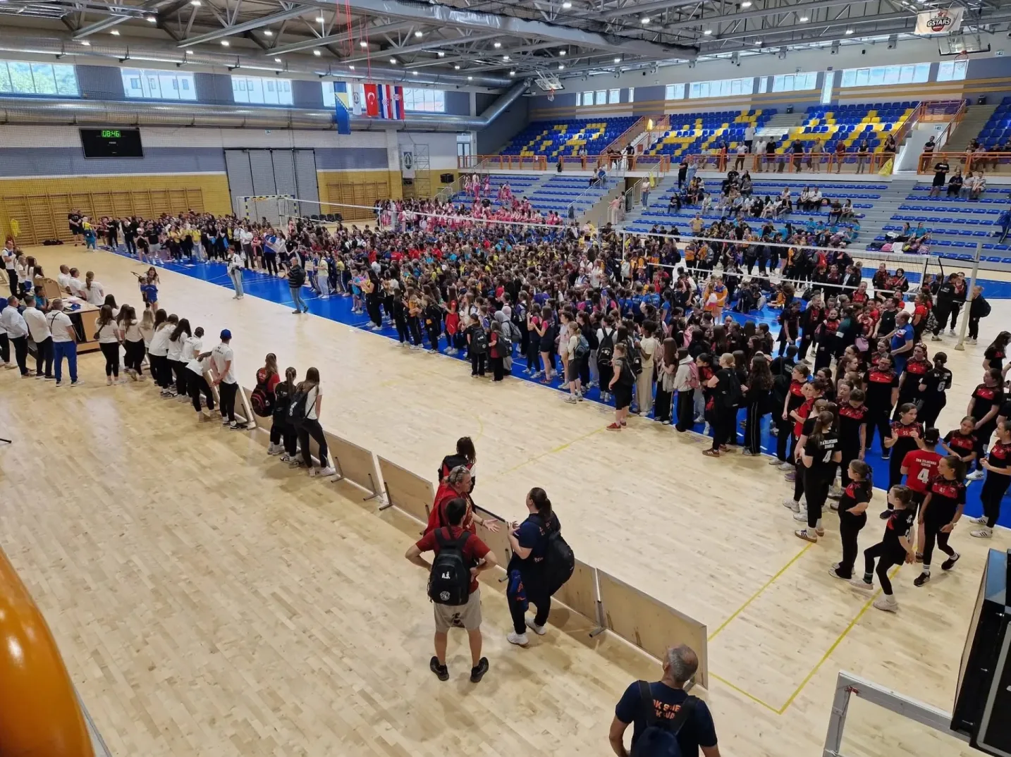 Veliki broj djece i odraslih u sportskoj dvorani, okupljeni na parketu i tribinama, vjerovatno na odbojkaškom takmičenju.