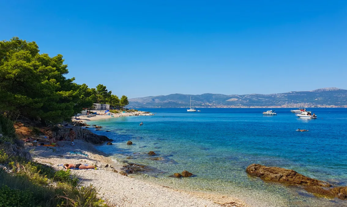 Plaža sa šljunkom i borovom šumom, ljudi se sunčaju, a u moru plove jahte i brodice, sa planinama u pozadini.