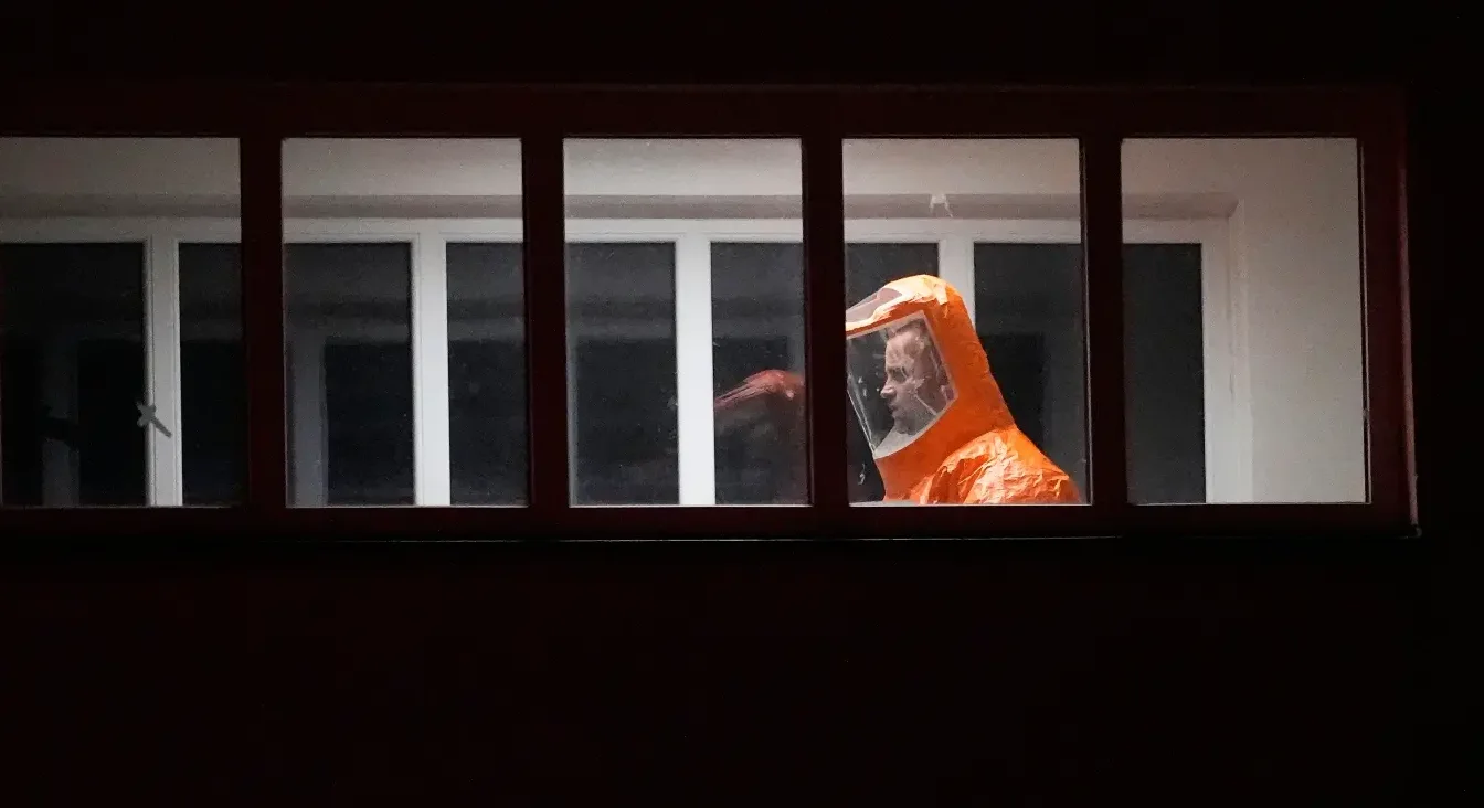 Osoba u narandžastom zaštitnom odijelu sa providnom kacigom gleda kroz mračni prozor.