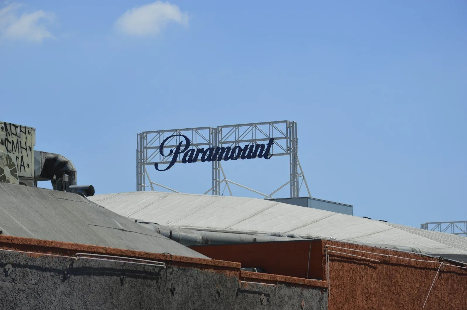 Plavi natpis "Paramount" ispisan elegantnim fontom, postavljen na metalnoj konstrukciji iznad zgrade pod vedrim plavim nebom.