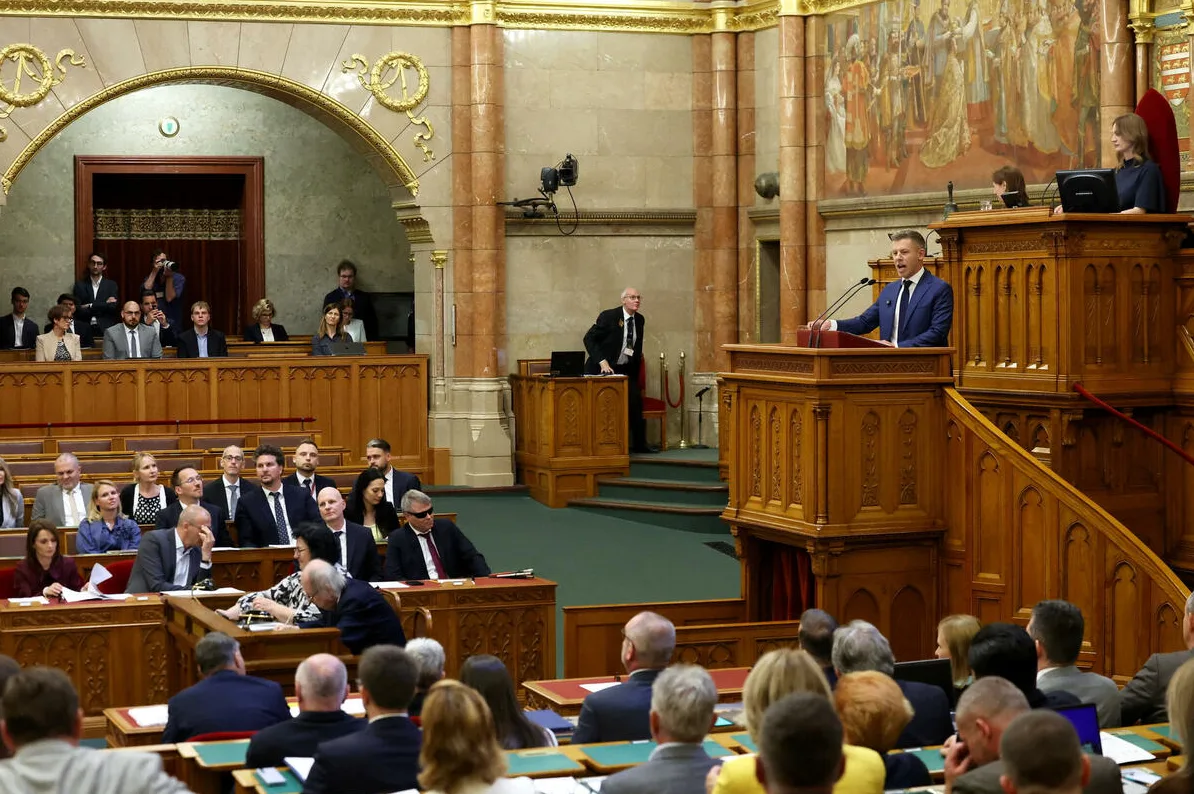 Političar u plavom odijelu govori za govornicom u parlamentu, okružen poslanicima koji sjede u klupama.