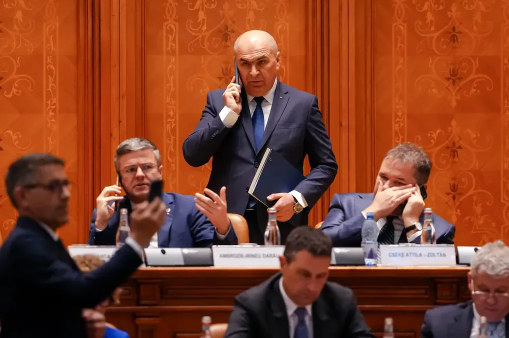 Grupa muškaraca u odijelima sjedi za stolom u sali, neki od njih razgovaraju telefonom, a jedan djeluje zabrinuto.