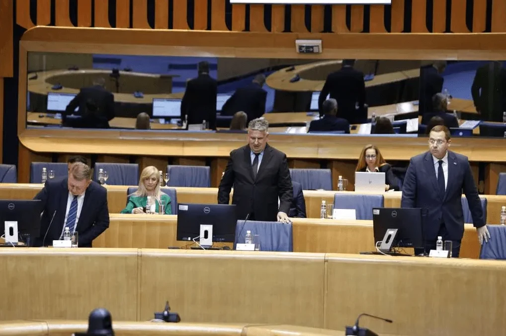 Grupa političara na sastanku u svecanoj sali, jedan govornik stoji za govornicom.