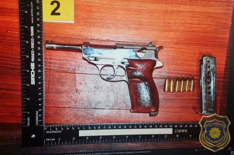 Prikazan je pištolj marke Walther P38 sa drvenim rukohvatom, zajedno sa pripadajućim metcima i okvirom. Pored predmeta nalazi se skala za mjerenje i oznaka '2'.