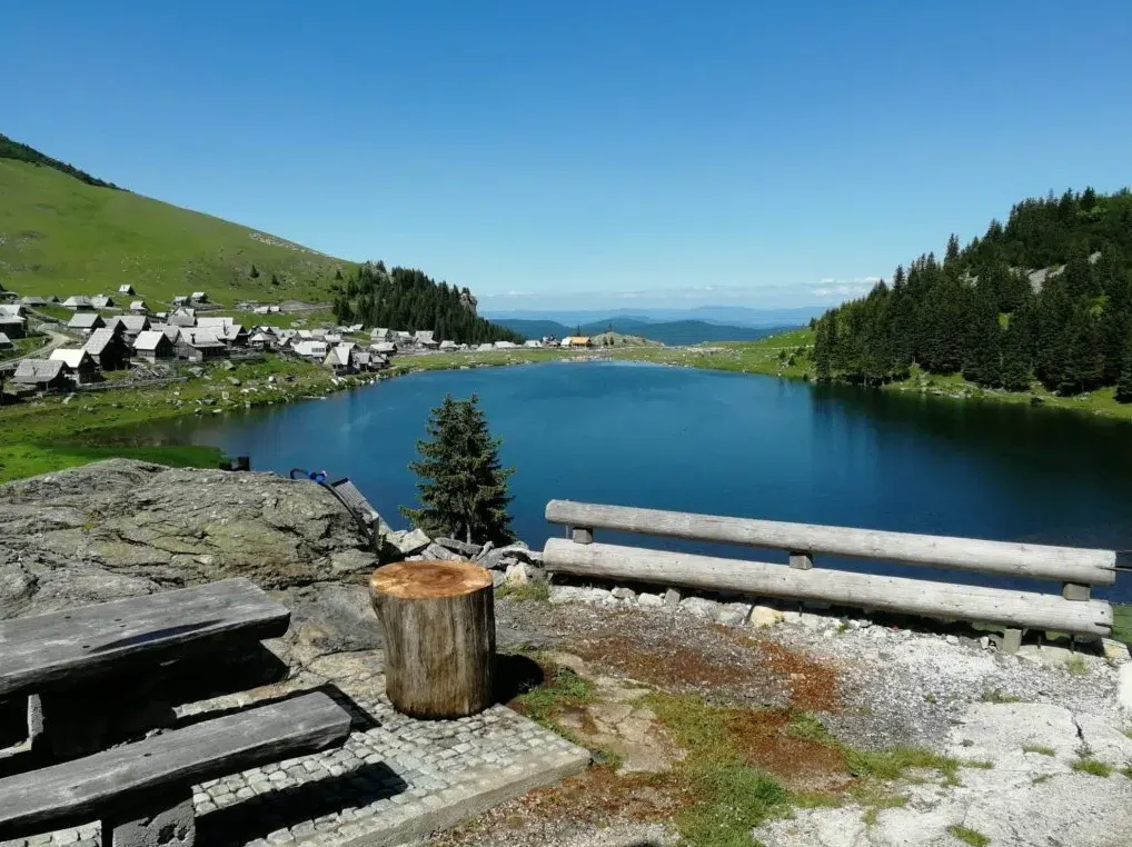 Mirno planinsko jezero okruženo zelenim obroncima, s malim drvenim kućama koje se nižu uz obalu i gustim borovima na obližnjim padinama.