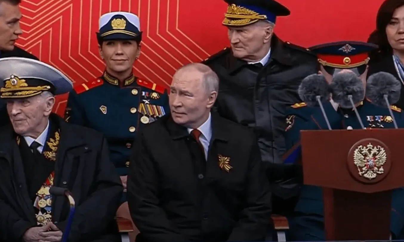 Vladimir Putin sjedi pored starijih muškaraca u vojnim uniformama i žena u uniformi, vjerovatno na vojnoj paradi u Moskvi.
