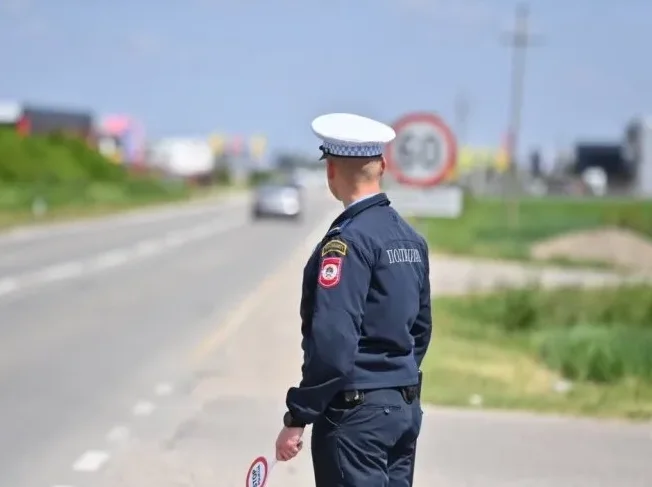 Policajac u uniformi saobraćajne policije drži "stop" znak dok stoji pored ceste sa ograničenjem brzine od 60 km/h u pozadini.