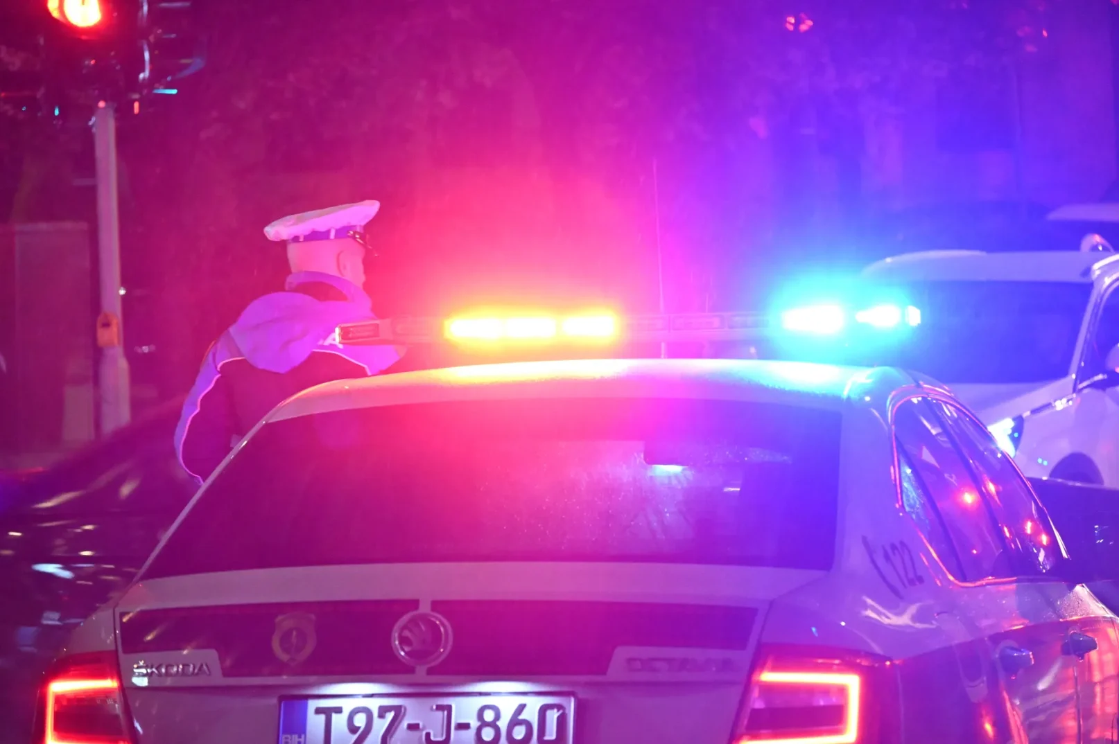 Policijski službenik u uniformi stoji pored službenog vozila s upaljenom rotacijom i trepćućim svjetlima u crvenoj, plavoj i žutoj boji.