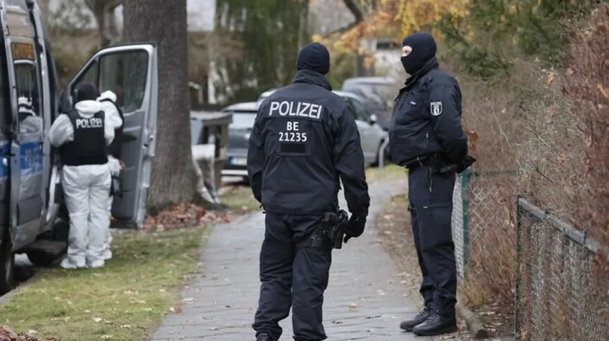 Dva policajca u uniformama stoje na trotoaru ispred kombija, dok treći policajac radi na otvorenim vratima vozila.
