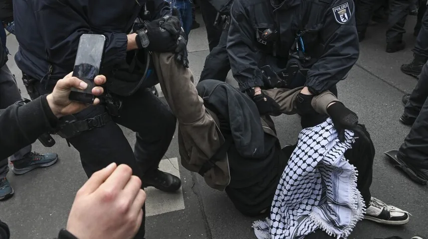 Policajci u uniformama odvode osobu na ulici, dok jedna osoba u prvom planu snima događaj pametnim telefonom.