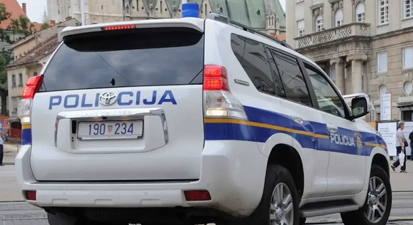 Bijeli policijski automobil marke Toyota Land Cruiser sa plavom rotacijom na krovu, parkiran na ulici u gradu. Na vozilu su ispisane oznake 'POLICIJA' plavim slovima, te plave i žute trake duž karoserije.