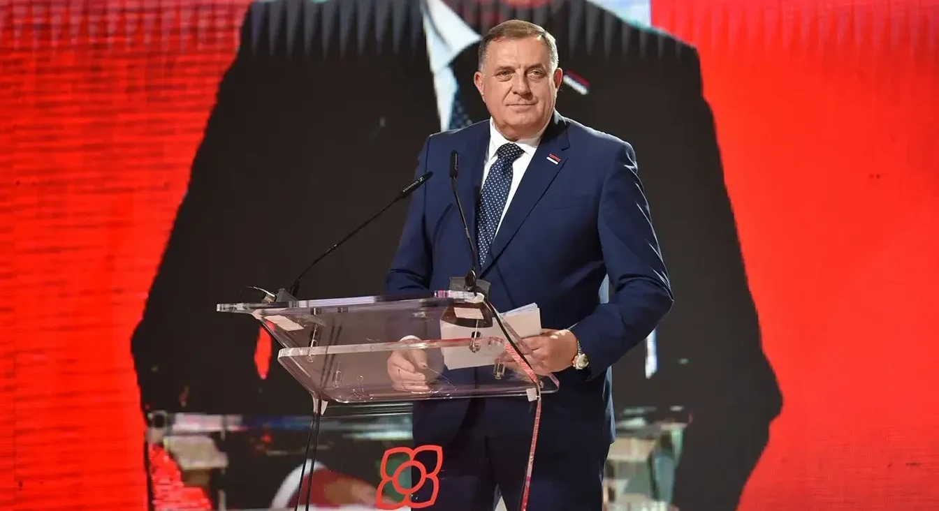 Političar u plavom odijelu stoji za govornicom s mikrofonima, držeći papire u ruci, ispred crvene pozadine sa velikim osvijetljenim likom u pozadini.