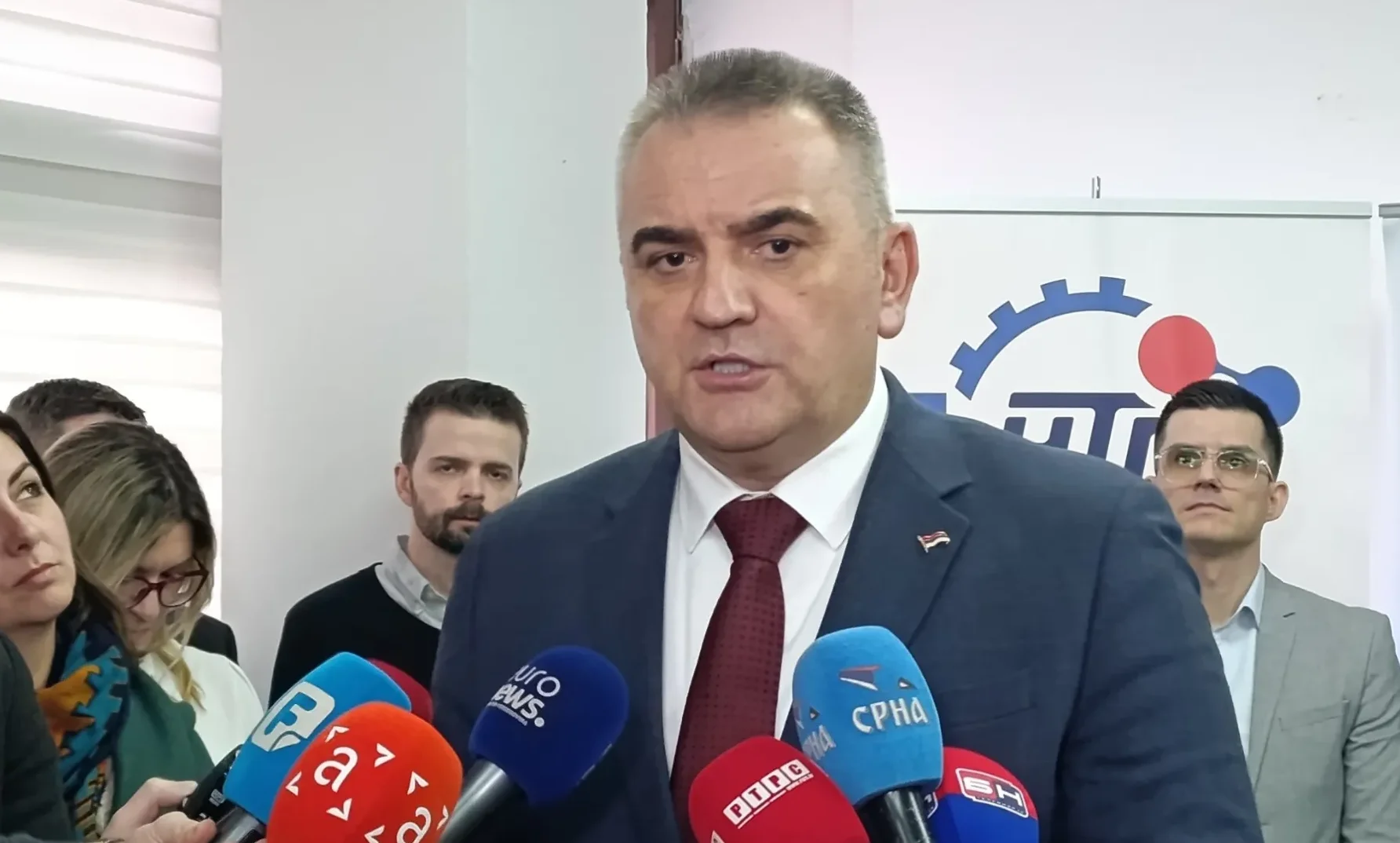 Političar u odijelu sa crvenom kravatom govori pred brojnim mikrofonima medijskih kuća.